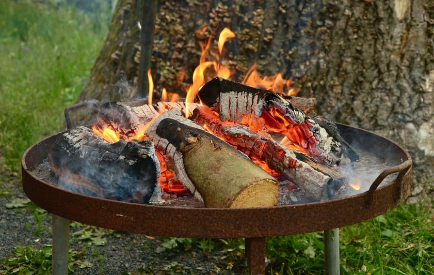 Feuerschale aus Cortenstahl mit brennendem Holz auf Kiesuntergrund neben Baum